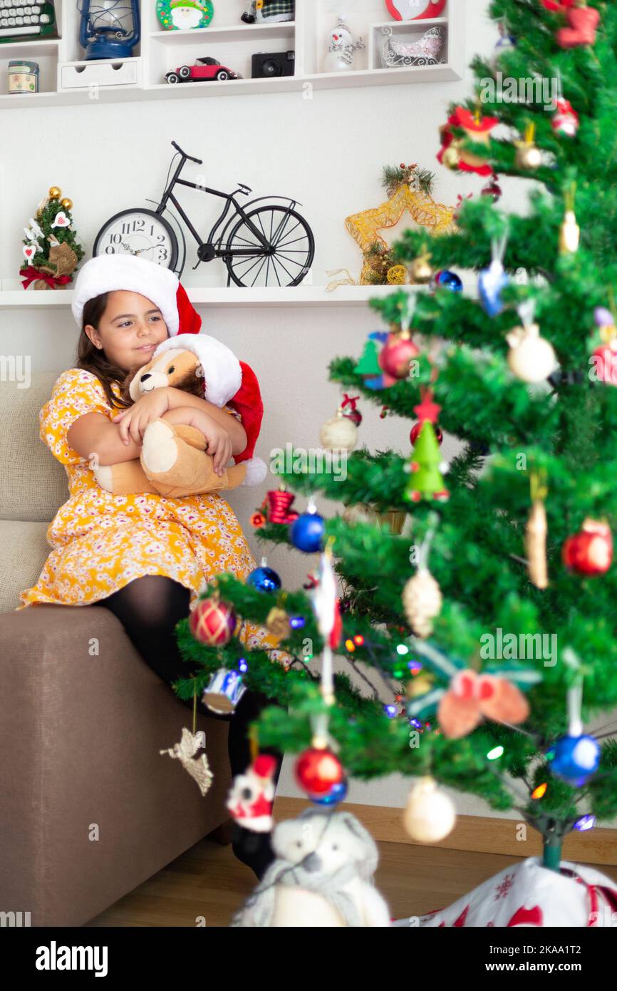 Petite fille embrassant son ours en peluche regardant l'arbre de Noël à la maison. Espace pour le texte. Banque D'Images