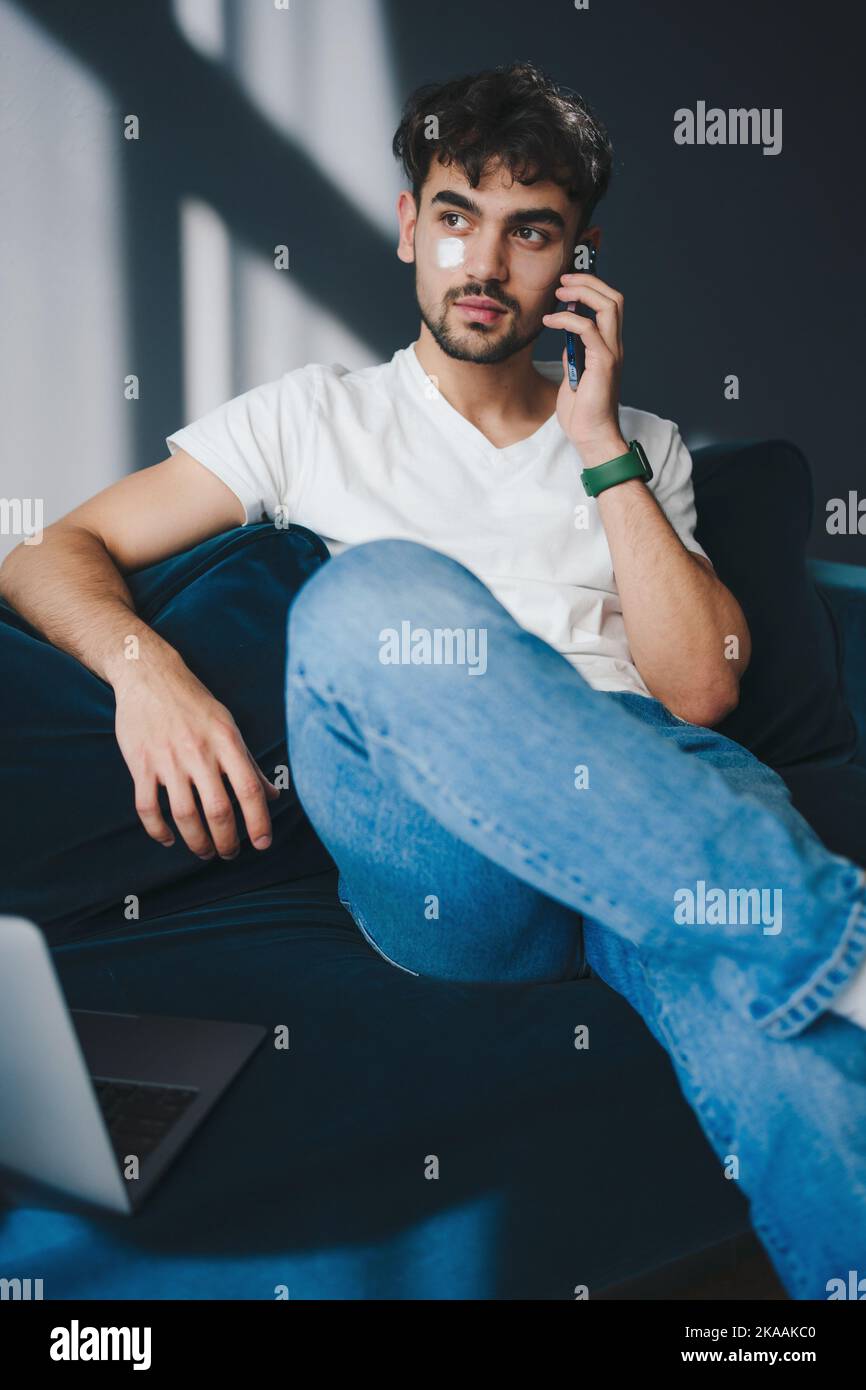 Homme caucasien assis sur un canapé bleu avec des taches d'hydrogel sous les yeux et travaillant à l'aide d'un ordinateur portable, ayant la conversation par téléphone. Routine de travail quotidienne Banque D'Images