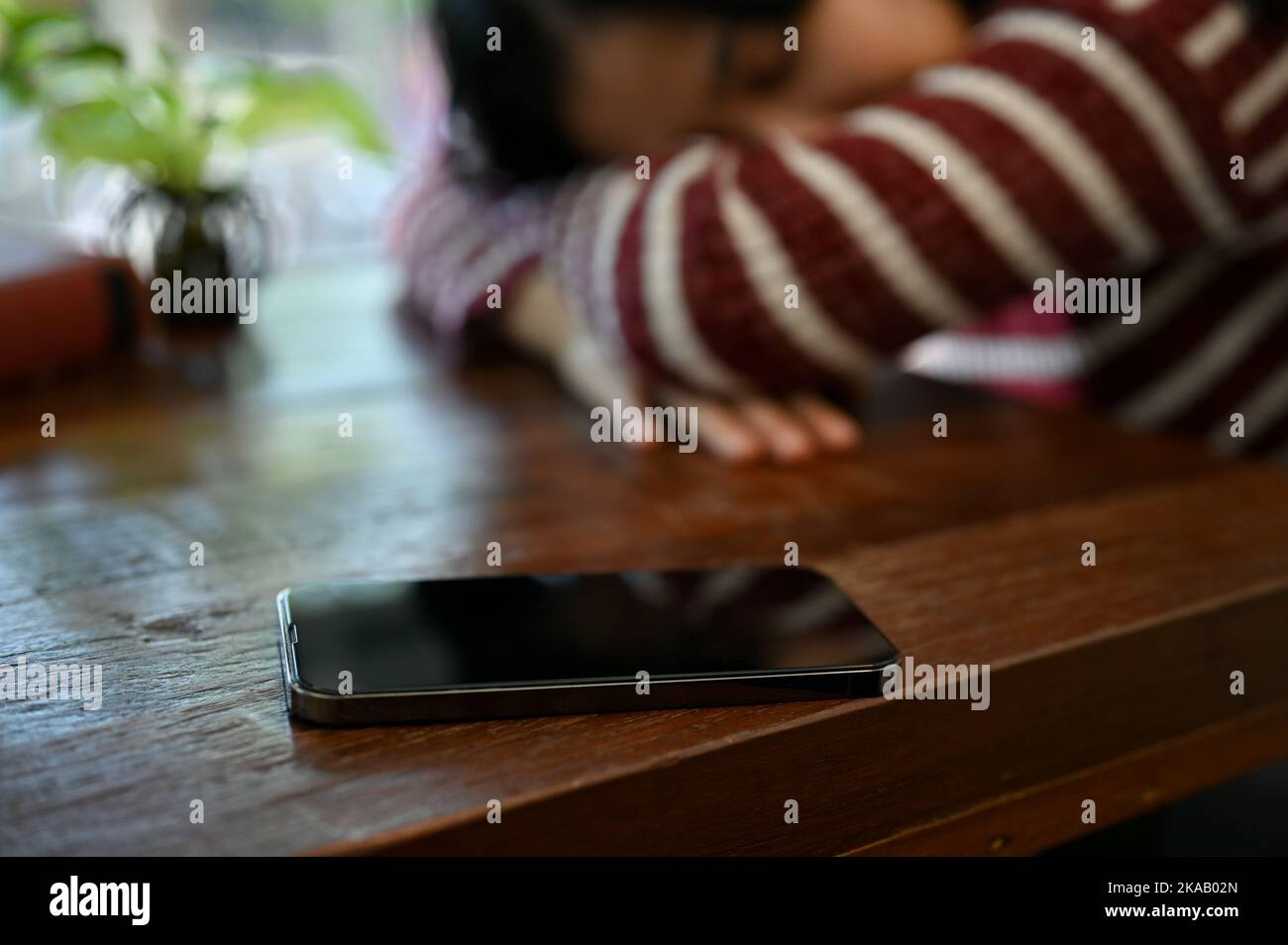 Un smartphone se trouve sur une table en bois sur un fond flou de jeune femme asiatique fatiguée qui la pose la tête ou qui la met à la table. Image de mise au point sélective Banque D'Images