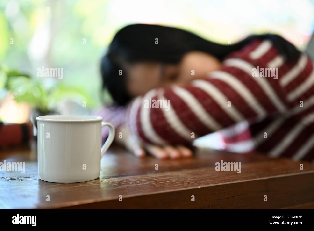 Une tasse de café blanc est sur une table en bois sur un fond flou de jeune femme asiatique fatiguée couché la tête ou se napper sur la table. Foyer sélectif Banque D'Images