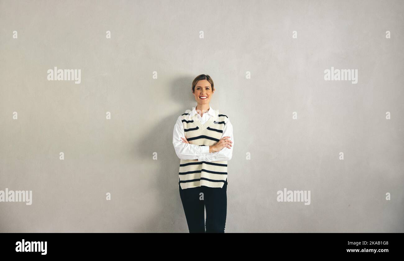 Bonne femme d'affaires souriant à la caméra tout en se tenant contre un mur gris avec ses bras croisés. Femme d'affaires gaie vêtue d'une tenue d'affaires décontractée Banque D'Images
