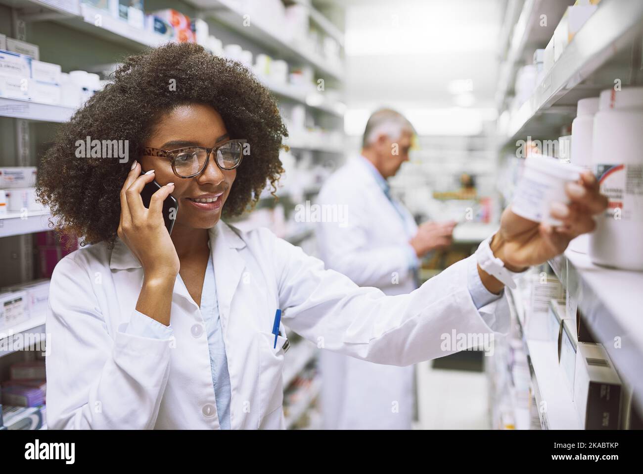 Nous avons effectivement ce médicament ici. Une jeune pharmacienne gaie qui parle sur son téléphone portable tout en lisant le dos d'une boîte de médicaments. Banque D'Images