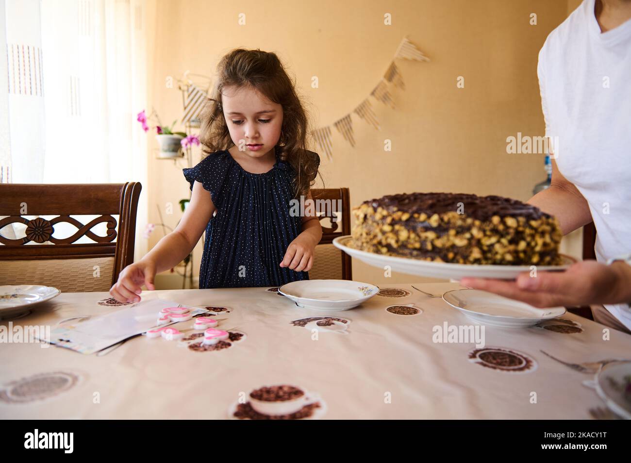 Charmante fille d'anniversaire en robe marine tendance, joue avec des bougies tandis que sa mère apporte un gâteau de chocolat festif sur la table Banque D'Images