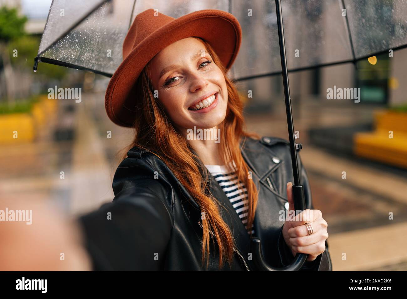 Jeune femme souriante à tête rouge portant un chapeau élégant sous un parapluie transparent faisant des selfie photo regardant l'appareil photo, tenant le téléphone portable à la main. Banque D'Images