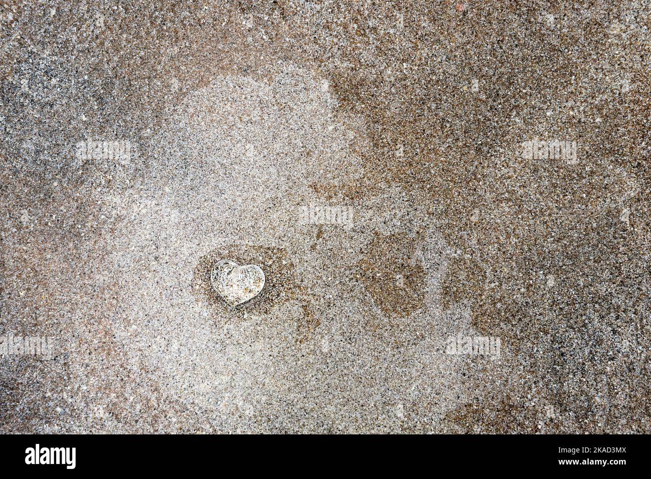 Petit coeur en verre clair sur le sable de la plage avec une fine couche d'eau. Banque D'Images
