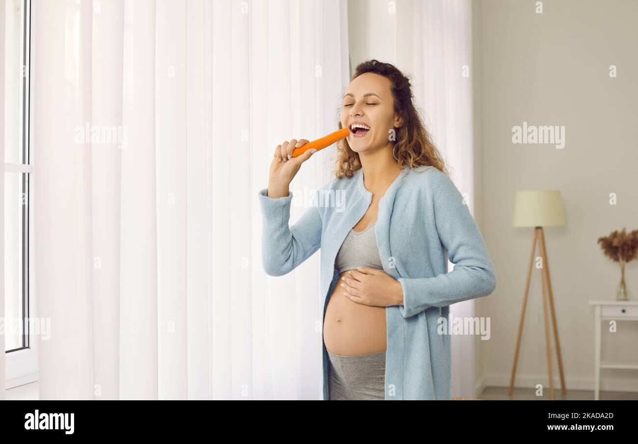 Bonne jeune femme enceinte souriante debout près de la fenêtre et mangeant une carotte fraîche Banque D'Images