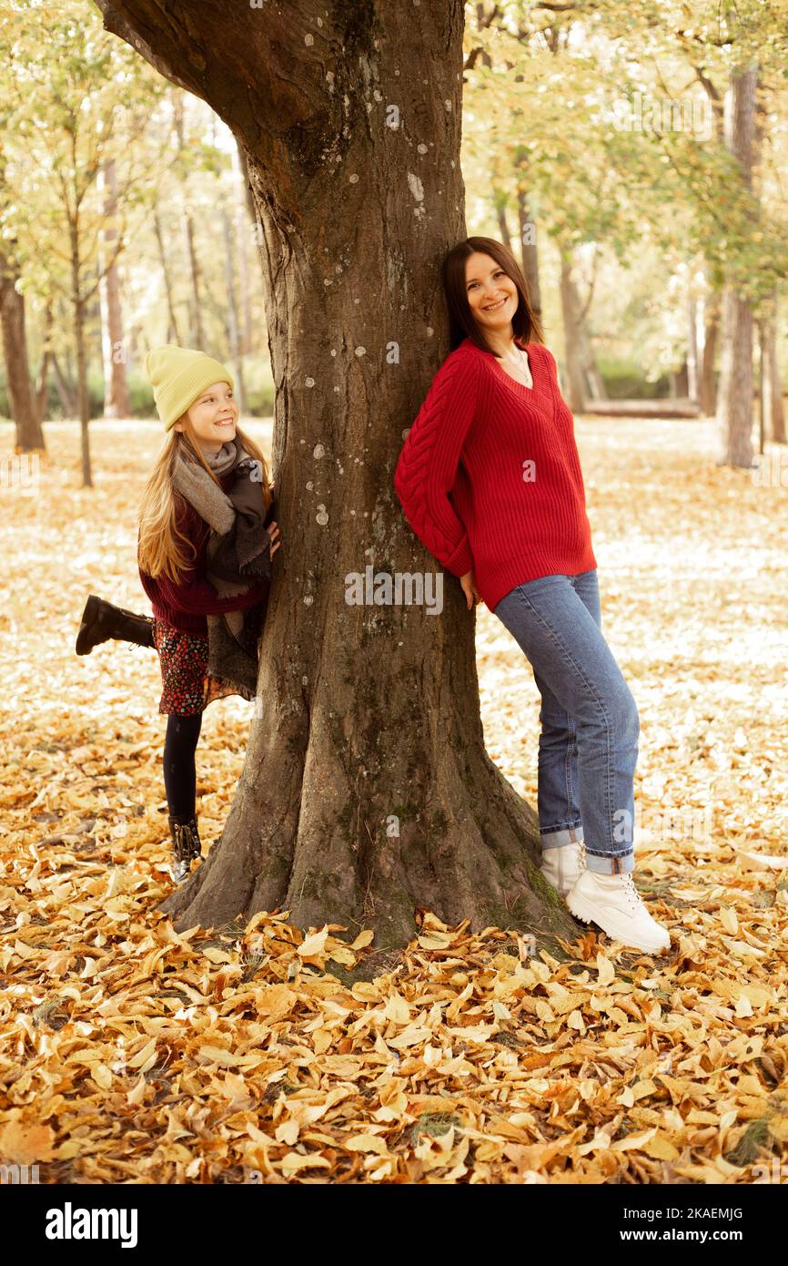 Photo pleine longueur de femme souriante et fille gaie jouant de cacher-et-chercher derrière l'arbre. Bonne mère et fille dans des tenues automnales douillettes ont le plaisir et Banque D'Images