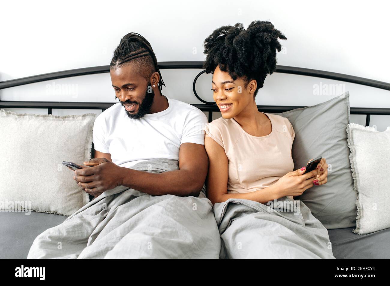 Joyeux couple afro-américain positif, heureux conjoints, femme et mari assis dans un lit confortable dans la chambre, utiliser leurs smartphones, la navigation sur les réseaux sociaux, l'actualité, la messagerie avec des amis, souriez Banque D'Images