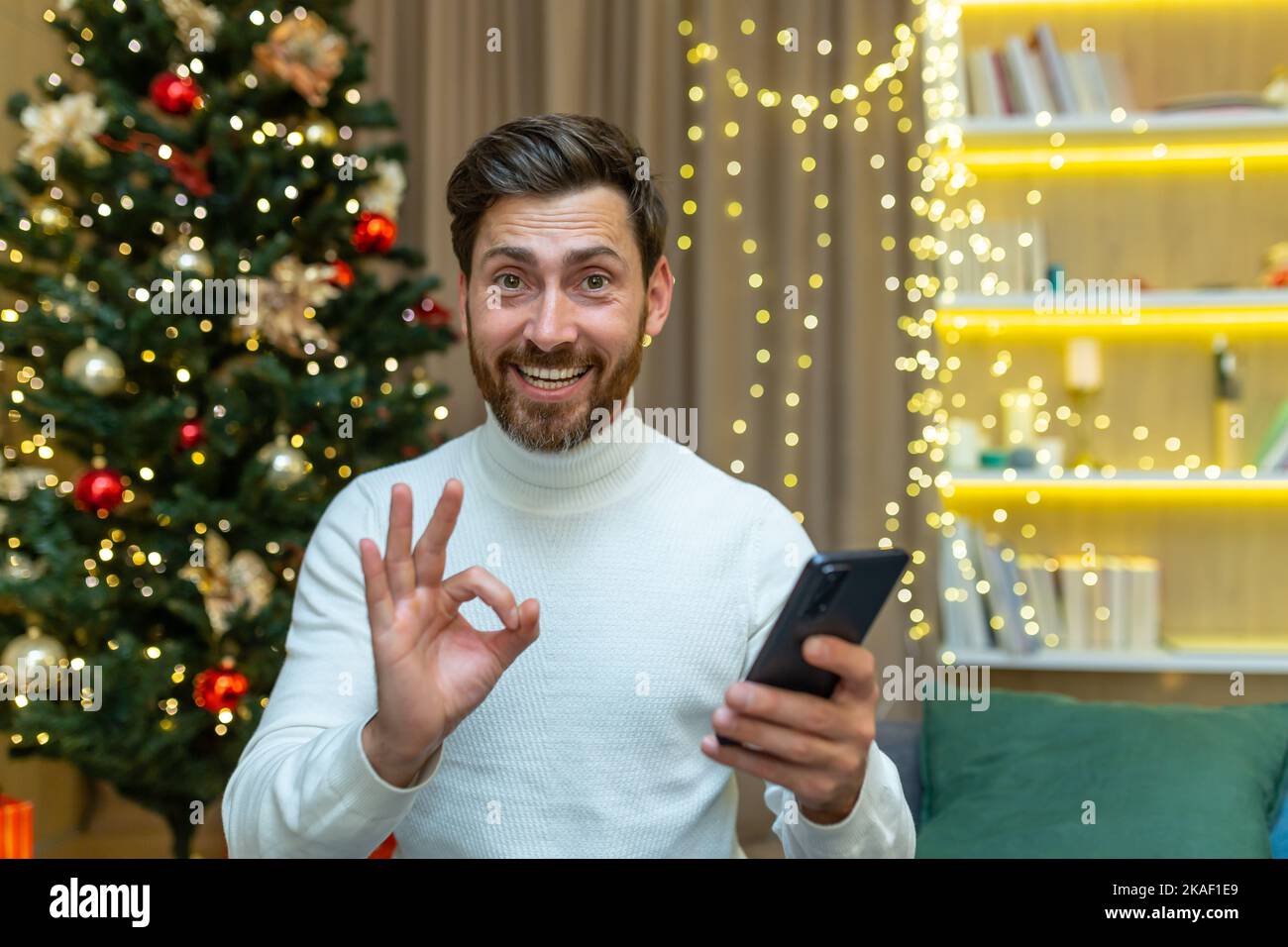 Achats en ligne festifs. Un jeune homme tient un téléphone entre ses mains, fait des achats en ligne, achète des cadeaux à des rabais de Noël, en vente. Il regarde la caméra, sourit, pointe avec ses doigts ok. Banque D'Images