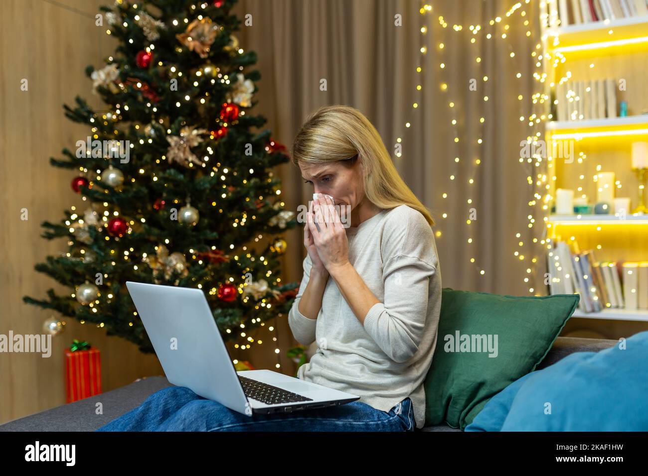 Une jeune femme est tombée malade pendant les vacances de Noël. Assis à la maison sur le canapé près de l'arbre, tenant un ordinateur portable, consultant avec un médecin par appel vidéo. Il essuie son nez avec une serviette, se sent mal. Banque D'Images