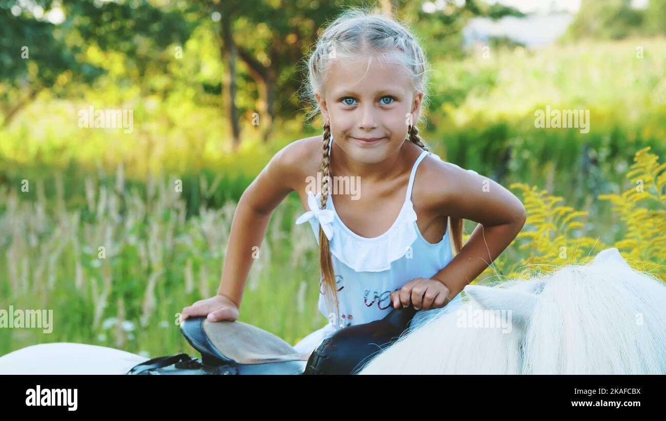 Blonde fille de sept ans, dans une robe blanche, monte, assis sur un poney. Bonnes vacances en famille. À l'extérieur, en été, près de la forêt. Photo de haute qualité Banque D'Images