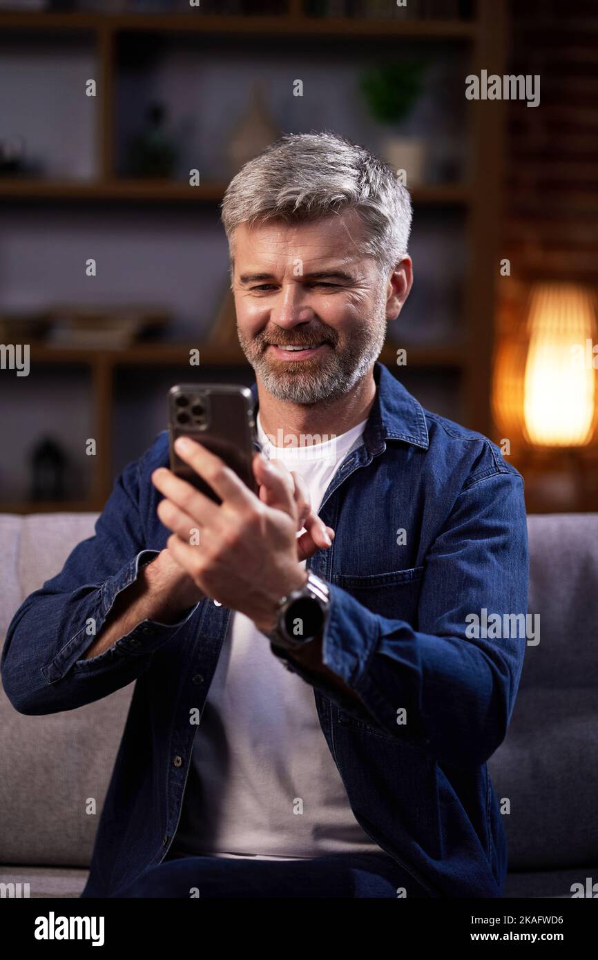 Homme souriant communique en ligne sur les réseaux sociaux et utilise un smartphone. Un homme en vêtements décontractés regarde à travers un téléphone mobile, communique dans un Banque D'Images
