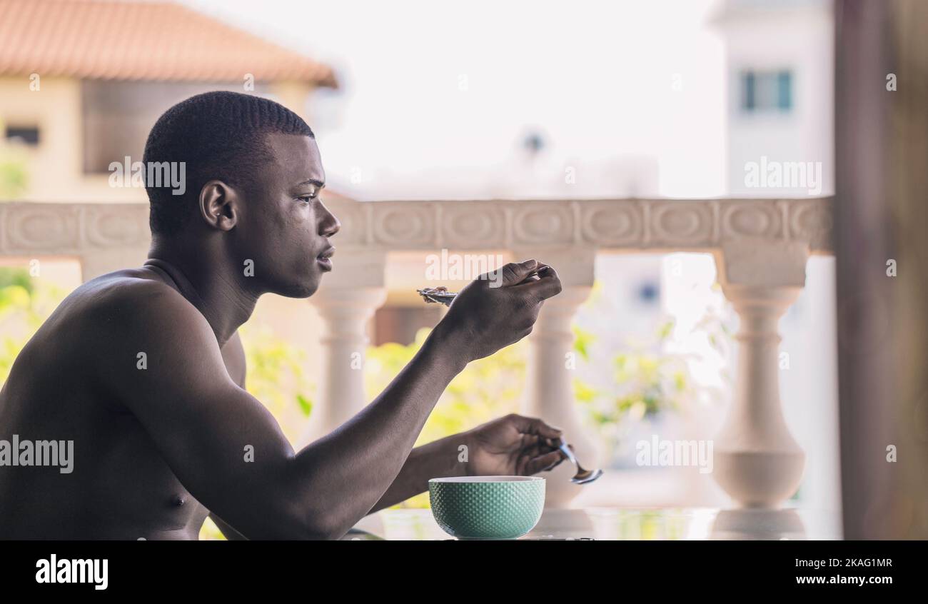 Vue latérale de l'homme afro-américain indéfable qui prend le petit-déjeuner tout en étant assis à table sur la terrasse de l'hôtel le matin Banque D'Images