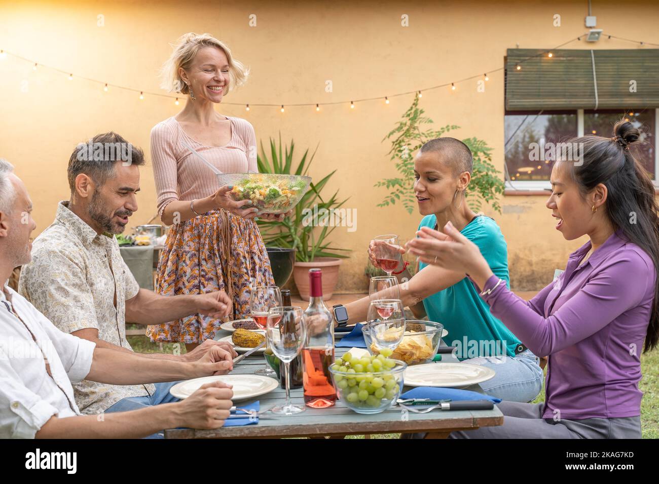 Des amis heureux rient avec un grand sourire autour de la table. La femme sert la nourriture dans le patio de la maison. Sourires, gaieté concept. Banque D'Images
