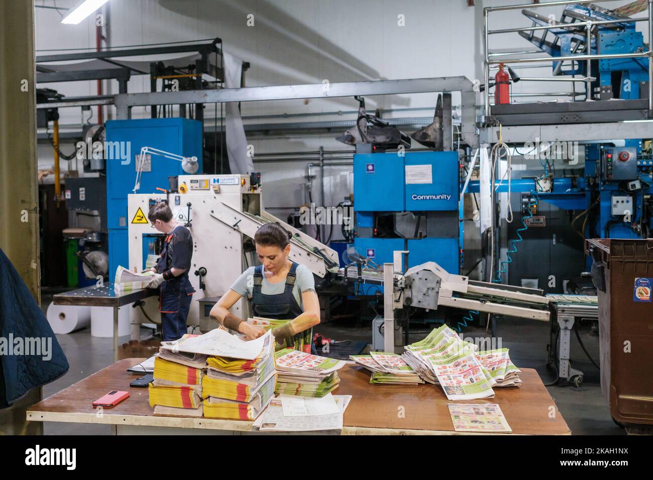 Vilnius, Lituanie - 12 octobre 2022: Femme travaillant en bloc d'empilage uniforme de magazines fraîchement imprimés sur table près de collègue et tapis roulant Banque D'Images
