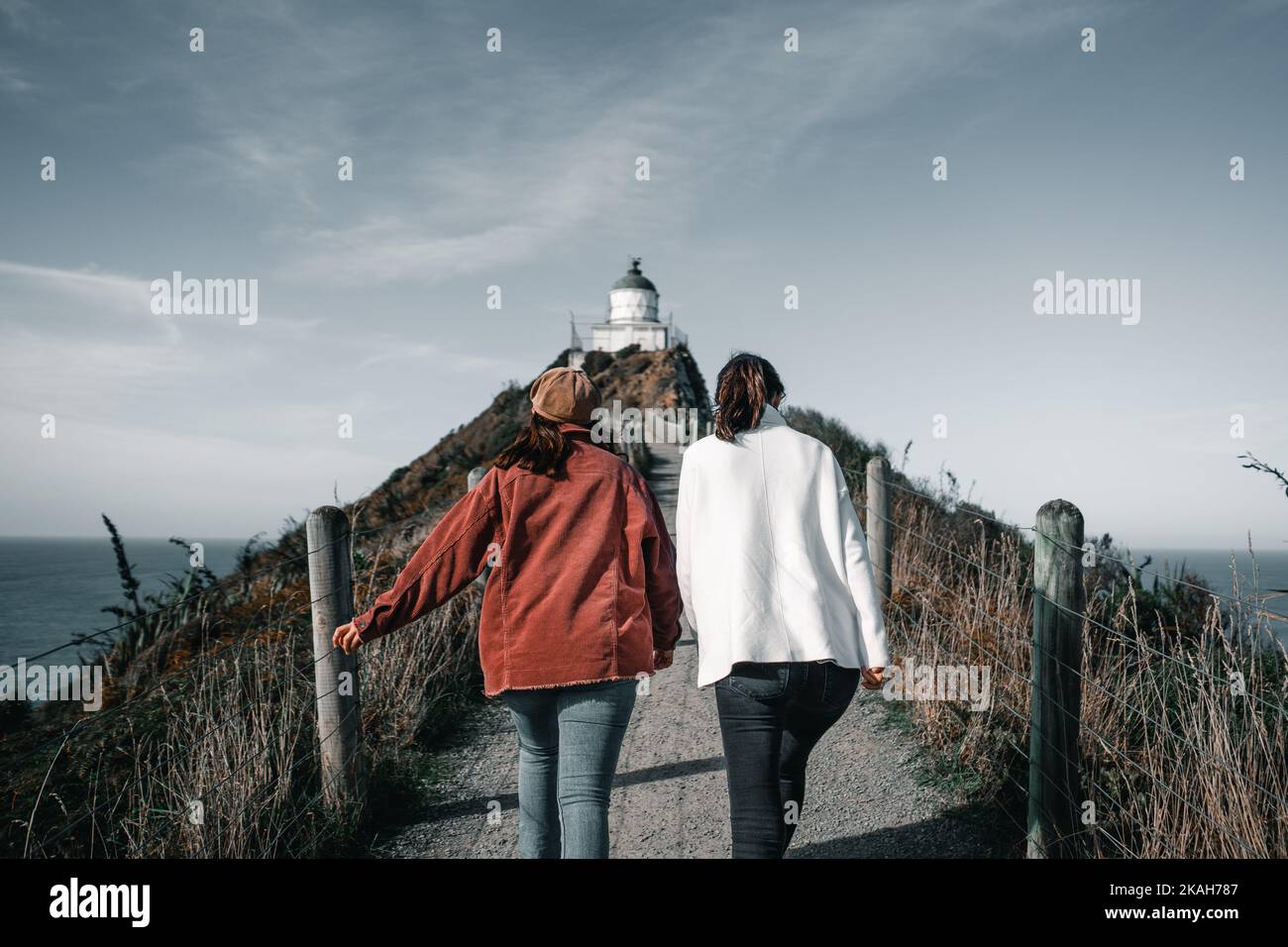 deux jeunes femmes caucasiennes marchant avec leur dos à la caméra vers le joli phare de sommet le long de la route de terre par la clôture sous un bleu Banque D'Images