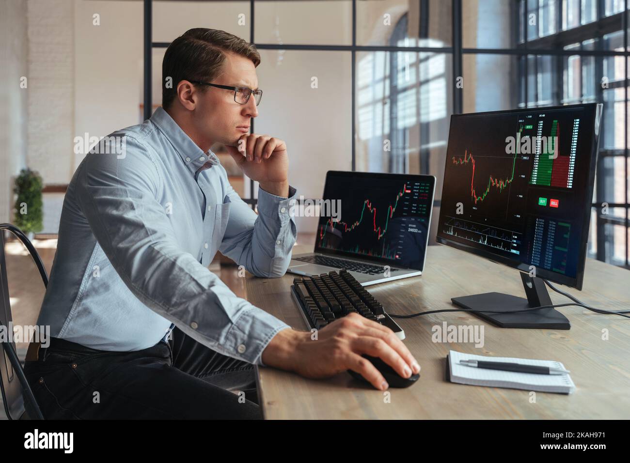 Vue latérale du broker crypto assis à la table du bureau, analyse du marché, regardant l'écran d'un ordinateur personnel avec un diagramme de chandeliers, touchant le menton avec la main, pensant, portant une chemise et des lunettes Banque D'Images