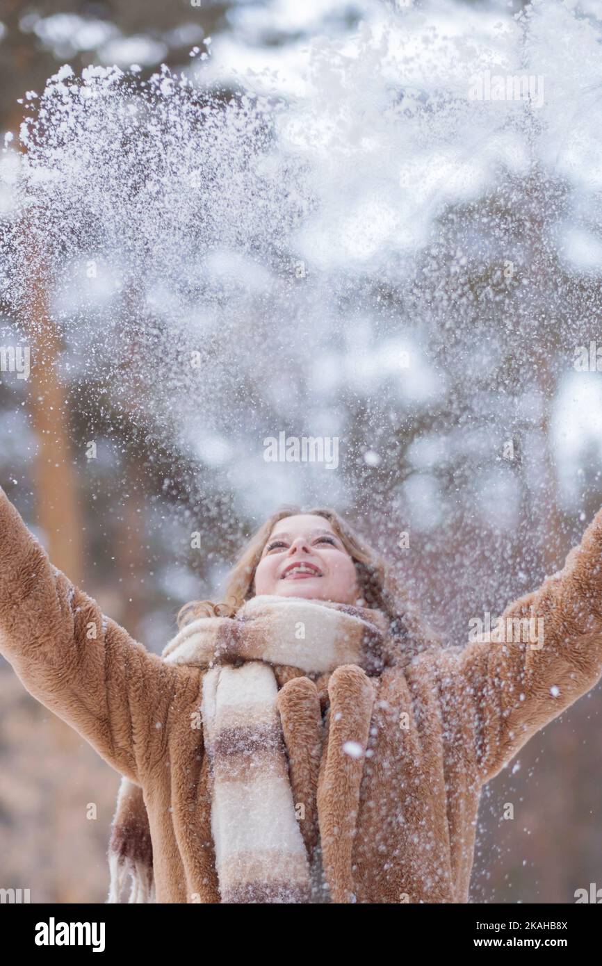 Une blonde blanche en bonnet tricoté, une longue écharpe et un manteau de  fourrure se promène dans la forêt et sourit. Femme attrape la neige dans  des moufles blanches. L'hiver est dans