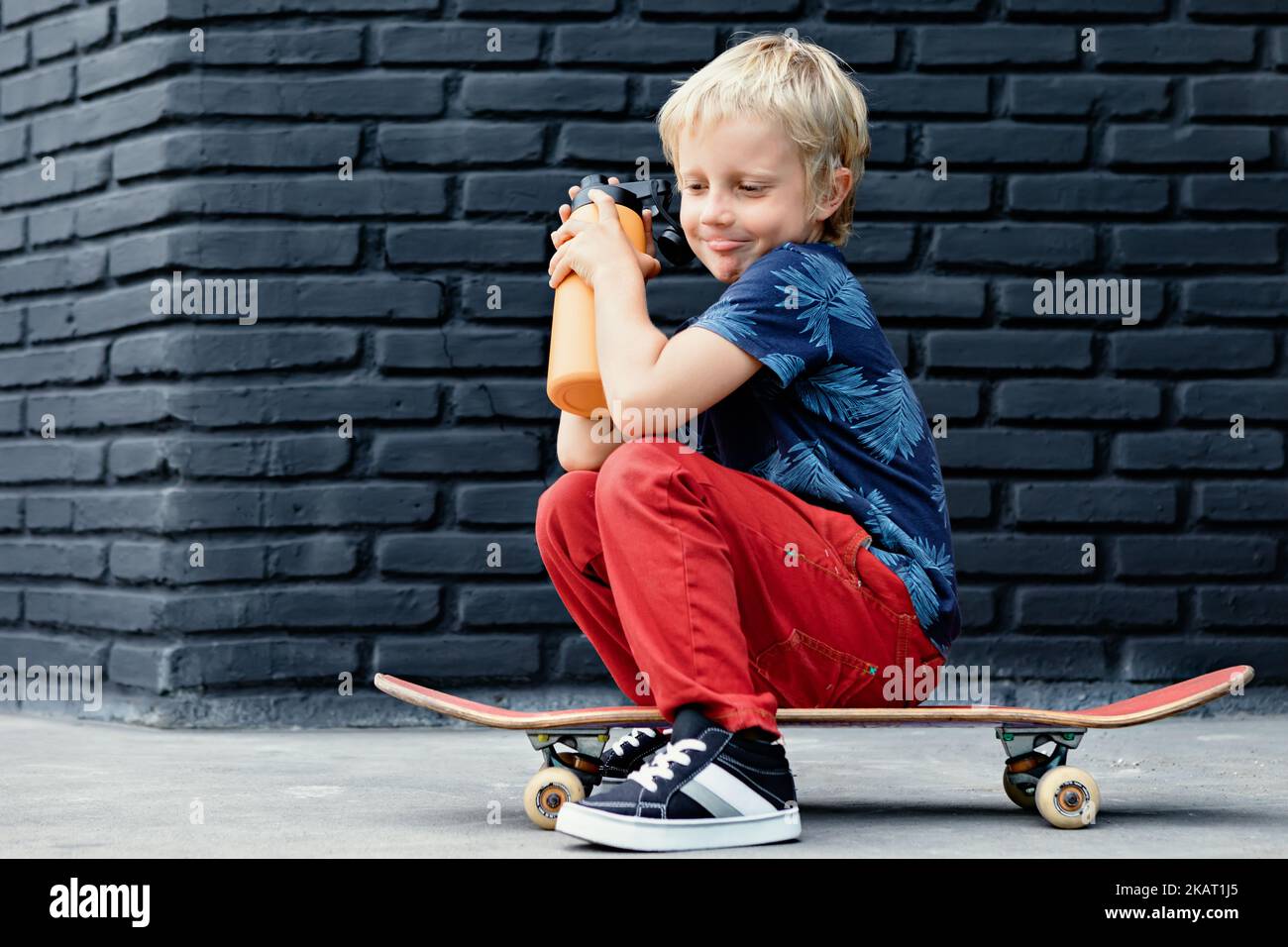 Le jeune patineur s'assoit sur le skateboard, boit de l'eau fraîche du biberon réutilisable après la formation des enfants. Vie familiale active, climatisation de loisirs en extérieur Banque D'Images