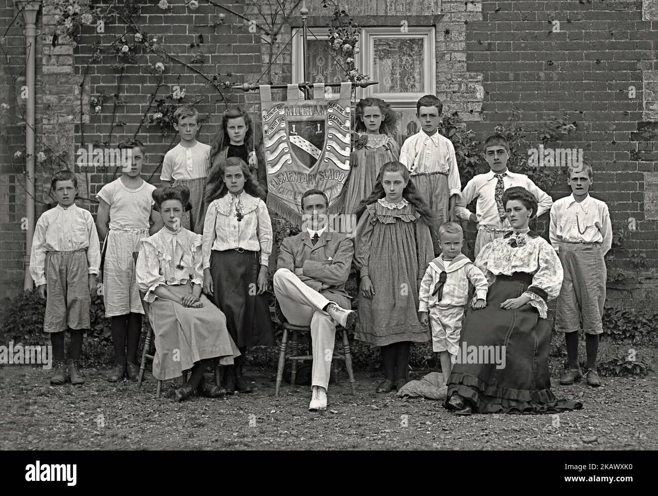 Une photographie de groupe d'élèves et de deux enseignants à l'école primaire Mileham, Breckland, Norfolk, Angleterre, R.-U. 1908. Les garçons et les filles portent un mélange intéressant de vêtements. Certains sont très habilement habillés, tout comme leurs professeurs. Certains des garçons semblent habillés pour les activités sportives. Ils sont posés avec une bannière avec le bouclier de l'école sur lui. Il indique également les gagnants de leur concours scolaire («trophée défi») – il semble que la classe Scarning a souvent remporté le prix! L'école a été fondée en 1677, mais elle a fermé en 2015, comme de nombreuses communautés rurales – une photographie victorienne/édouardienne d'époque. Banque D'Images