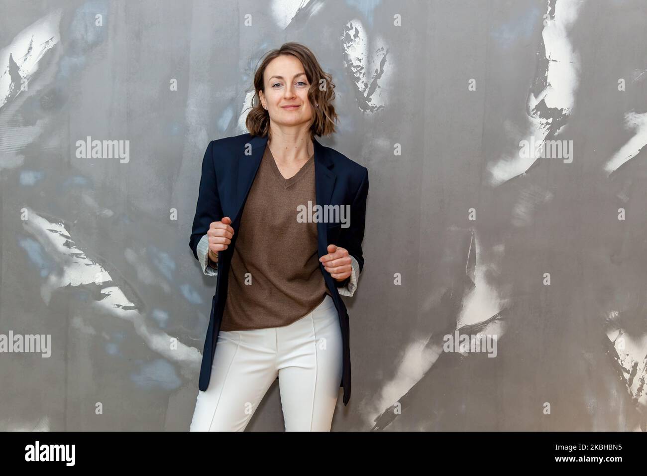 Tendance et élégante, elle pose dans un pantalon blanc, un pull marron et une veste noire contre un mur gris Banque D'Images