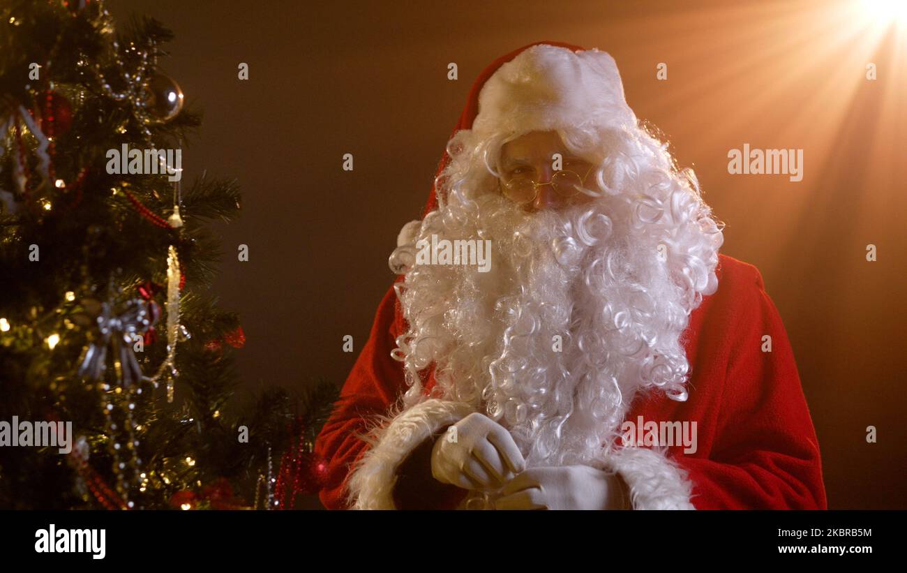 Le Père Noël porte un jouet du nouvel an sur l'arbre de Noël. Un homme habillé comme le Père Noël a l'air poupe. Le Père Noël est sur l'appareil photo et semble sterniquement. Le Père Noël Banque D'Images