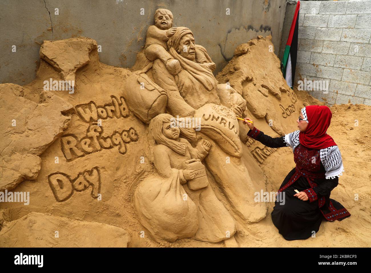 Le sculpteur de sable palestinien Rana Ramlawi termine une nouvelle création d'œuvres commémorant la Journée mondiale des réfugiés, journée consacrée par l'Assemblée générale des Nations Unies à la sensibilisation à la situation des réfugiés dans le monde entier, à Gaza, à 20 juin 2020, Illustration d'une femme portant un enfant et d'un sac de farine portant le logo de l'Office de secours et de travaux des Nations Unies pour les réfugiés de Palestine (UNRWA) avec un enfant qui tient une jerrican d'eau, tous devant une carte du monde. - Plus de 1,5 millions de personnes vivent dans 58 camps de réfugiés palestiniens reconnus en Jordanie, à Leban Banque D'Images