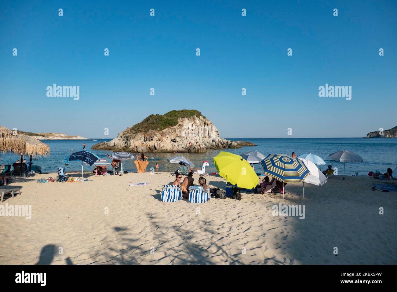 La petite île près de la rive de Kalamitsi. Foule de touristes à la célèbre et populaire plage de Kalamitsi située à la pointe sud de la péninsule de Sithonia à Chalkidiki dans le nord de la Grèce. Les gens s'amusent à la plage en Grèce car ils se détendent à l'ombre des parasols, plongée en apnée, plongée sous-marine, natation dans les eaux cristallines tropicales exotiques, sports nautiques, location de bateaux, prendre un verre dans les bars de la plage ou prendre un bain de soleil sur le sable doré et profiter de la vie sur la plage sous le ciel bleu ensoleillé. Il y a une petite île également située près de la rive. Le gouvernement local Banque D'Images