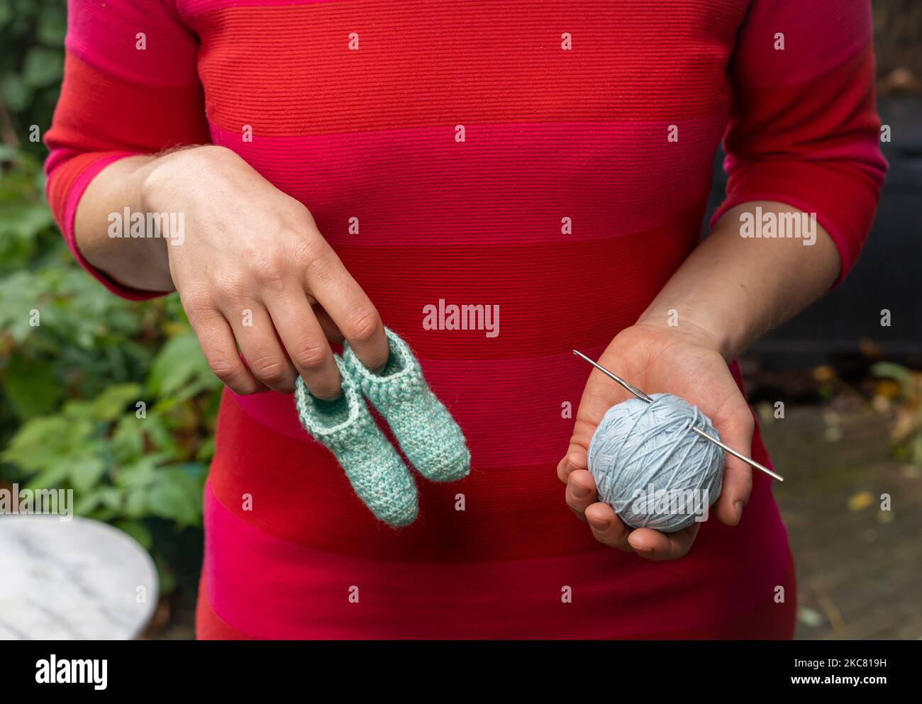 Femme en robe rouge tenant des chaussettes ou des chaussons faits main crochet bébé en laine d'une main, et boule de laine avec aiguille crochet dans l'autre, sélectionnez Banque D'Images