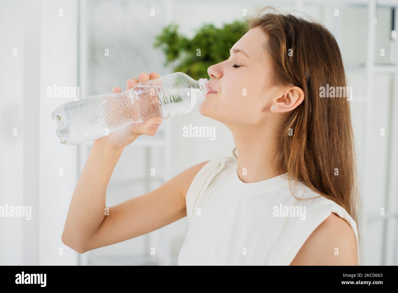 Jeune femme sportive une eau potable dans une bouteille en plastique debout par fenêtre à la maison, Santé, gens, nourriture, sports, concept de style de vie et de beauté Banque D'Images