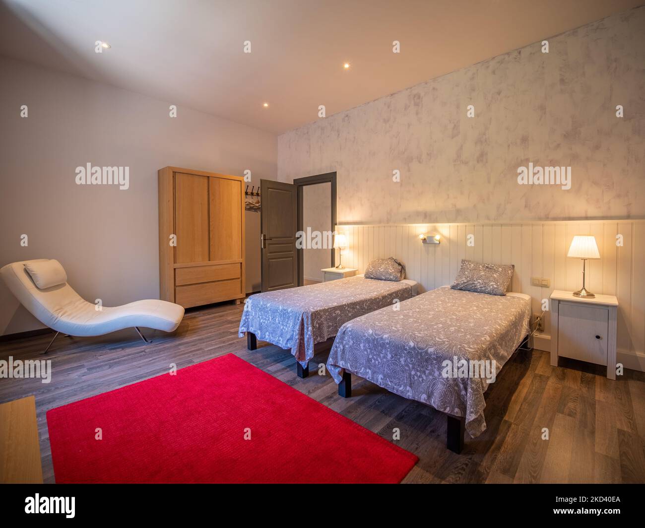 Une belle photo de chambre spacieuse avec deux lits simples, des murs clairs, du parquet, de la moquette rouge et un fauteuil Banque D'Images