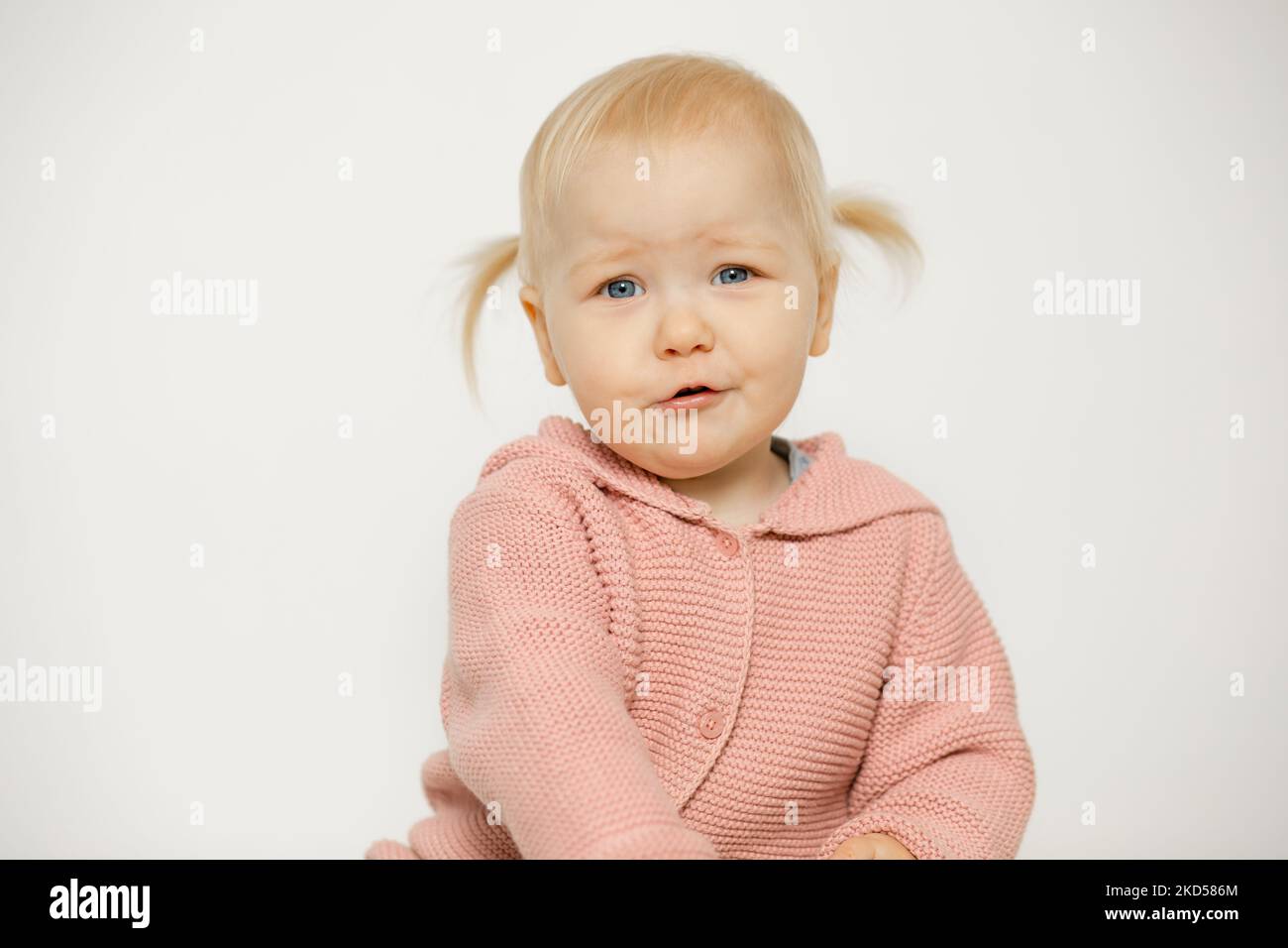 Petite fille malheureuse isolée sur blanc. Portrait d'un enfant triste en studio. Enfant blond aux cheveux avec une expression faciale incontentée en tricot rose Banque D'Images