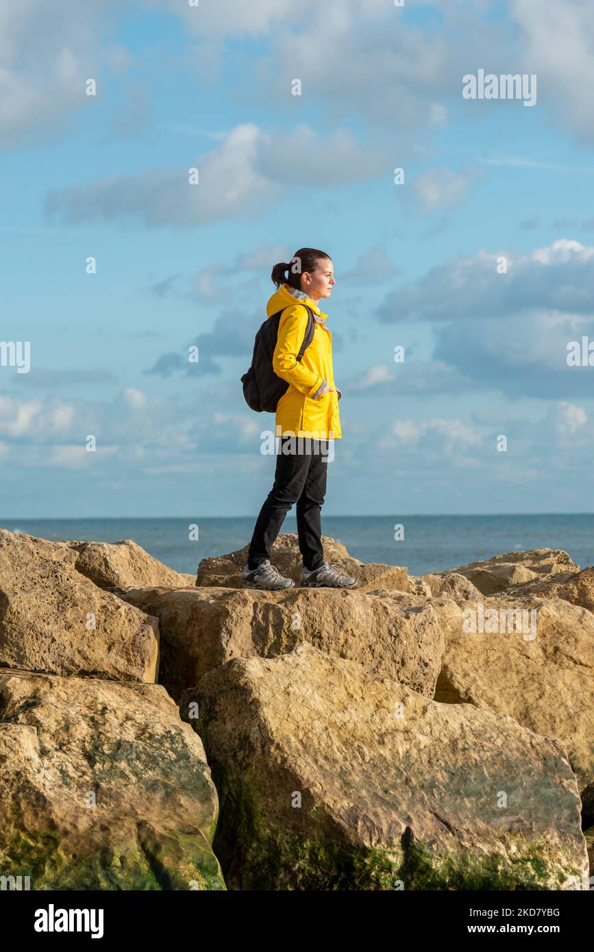 Femme portant un manteau jaune et un sac à dos marchant à travers les rochers au bord de la mer Banque D'Images