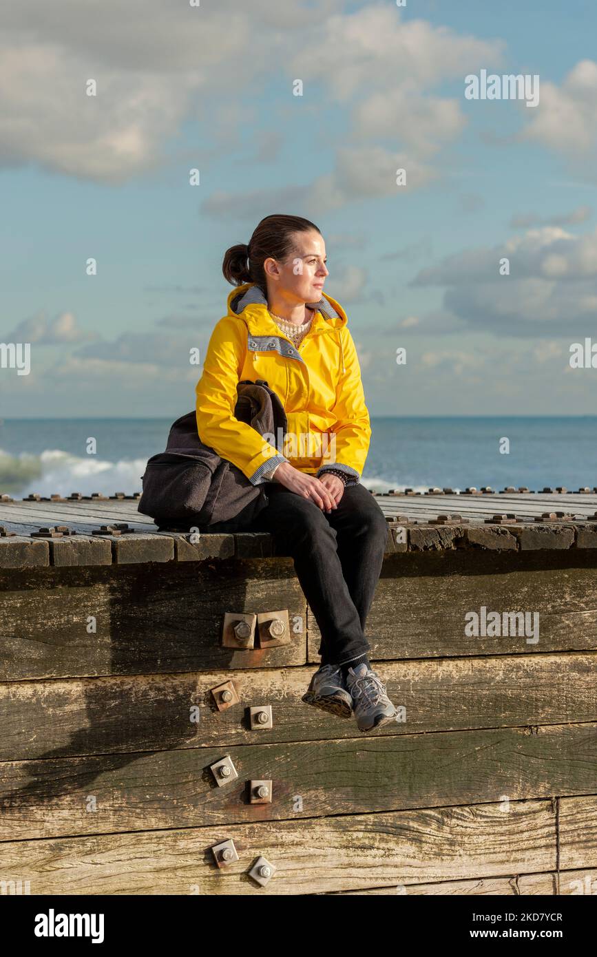 triste femme assise seule au bord de la mer, portant un manteau jaune. Banque D'Images