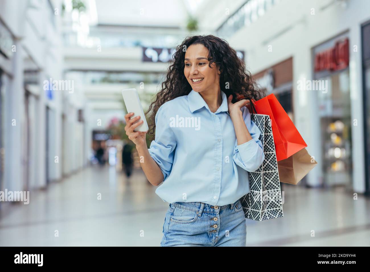 Une femme heureuse et prospère magasiner pour des vêtements dans un supermarché, une femme hispanique tenant un smartphone lisant des messages en ligne et parcourant des offres avec des réductions et des ventes Banque D'Images