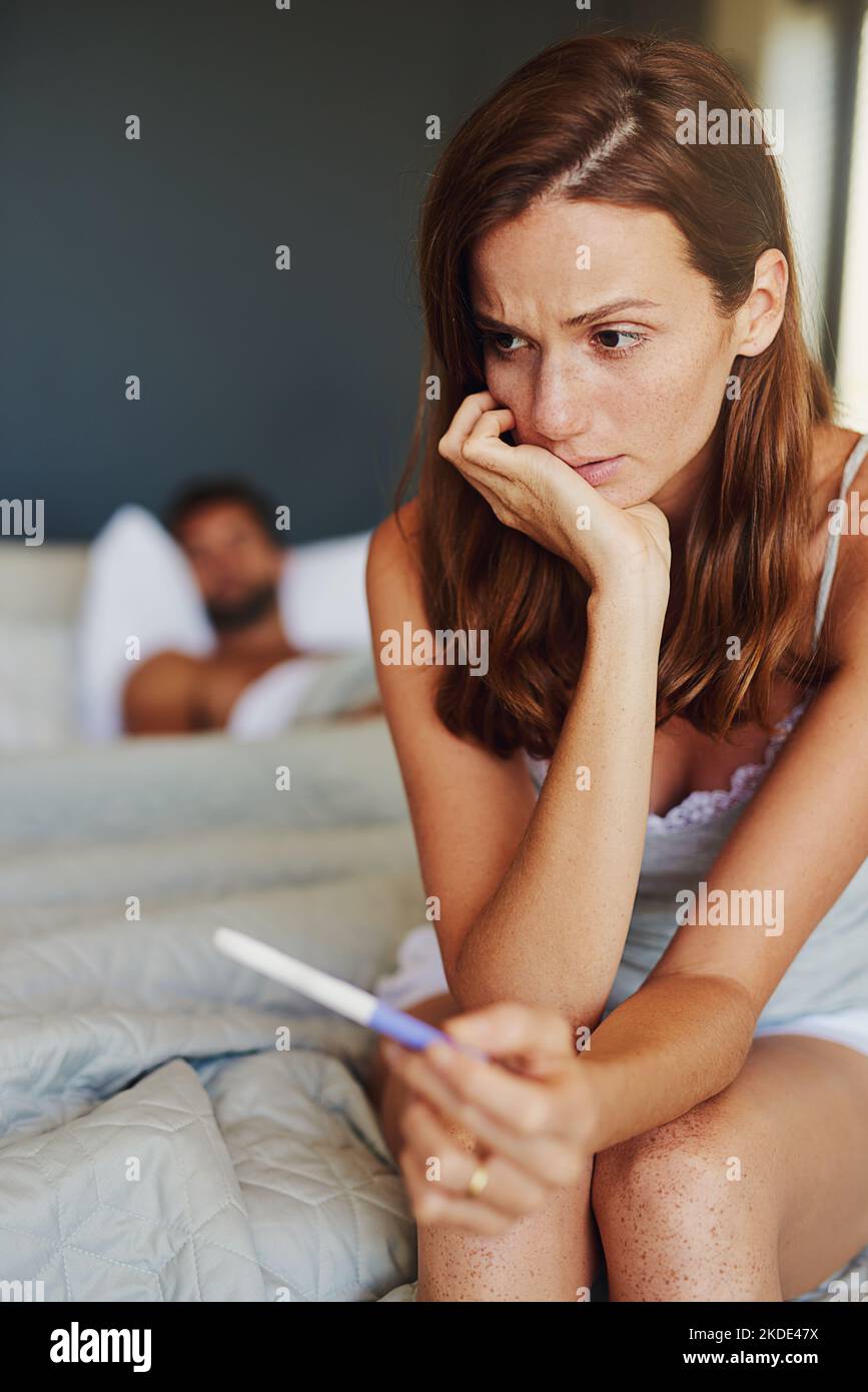 Comment va-t-il se sentir à ce sujet... une femme qui semble inquiète en tenant un test de grossesse avec son partenaire couché en arrière-plan. Banque D'Images