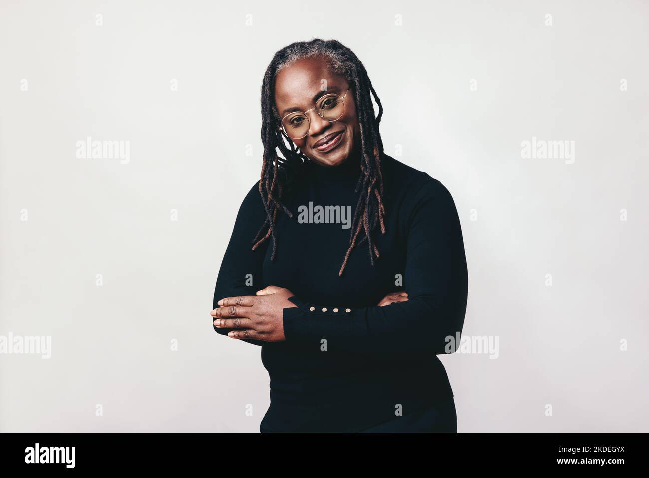 Femme professionnelle avec des dreadlocks souriant à l'appareil tout en portant des lunettes. Heureuse femme mûre debout sur fond gris avec ses bras Banque D'Images