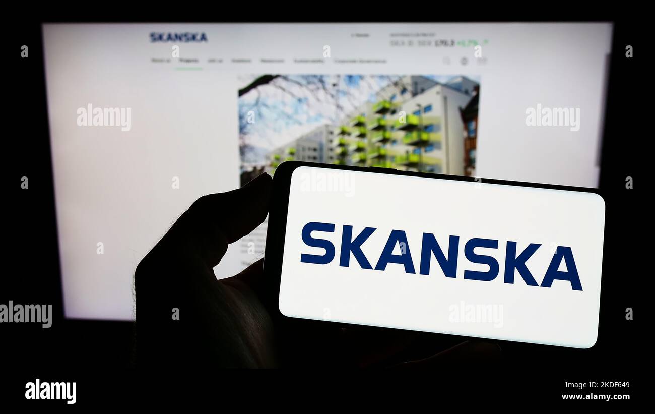 Personne tenant un téléphone portable avec le logo de la société suédoise de construction Skanska AB à l'écran en face de la page web d'affaires. Mise au point sur l'affichage du téléphone. Banque D'Images