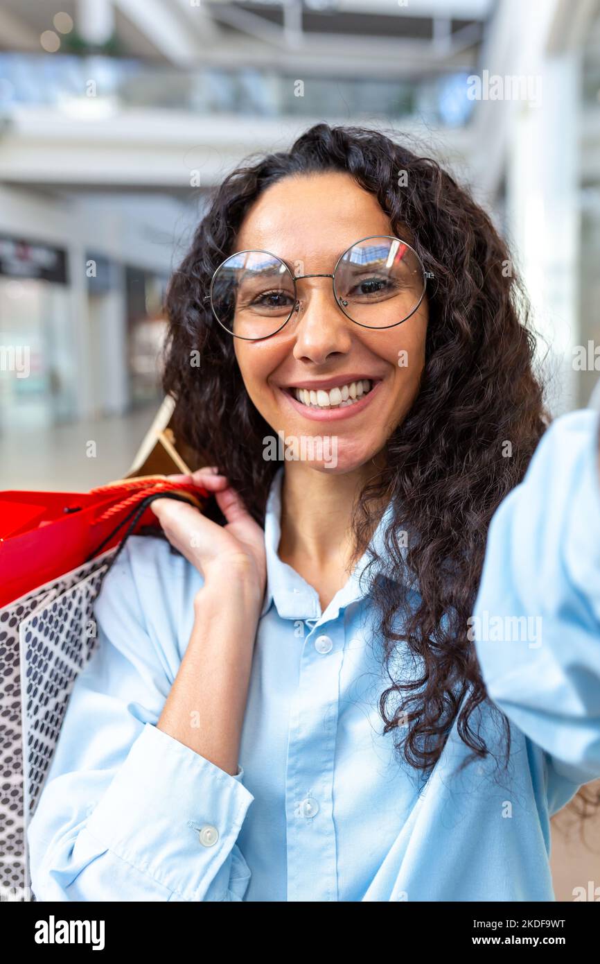 Belle femme hispanique regardant l'appareil photo de smartphone parler pendant un appel vidéo et prendre des photos de selfie, magasiner dans un grand magasin tenant des sacs-cadeaux colorés dans les mains souriant parler avec des amis. Banque D'Images