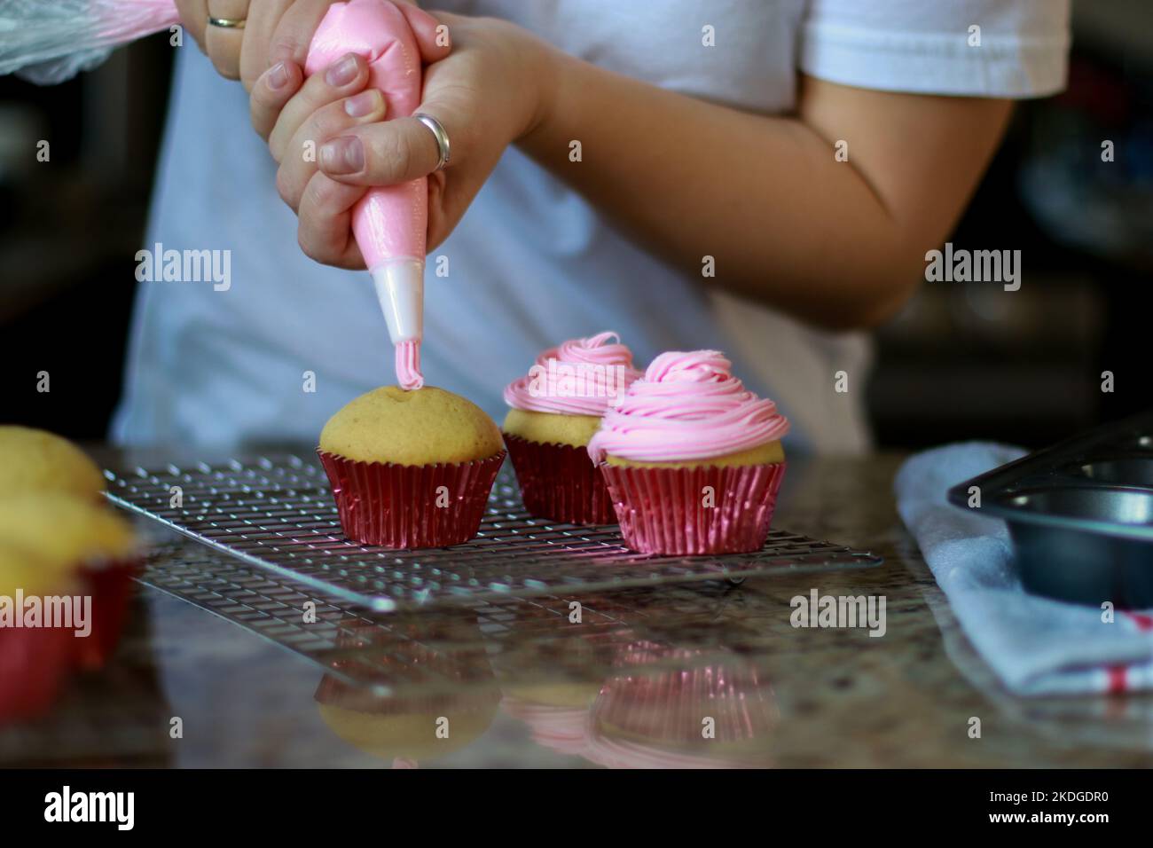 femme en chemise blanche étalant la crème de beurre rose givrant sur trois cupcakes, arrière-plan flou Banque D'Images