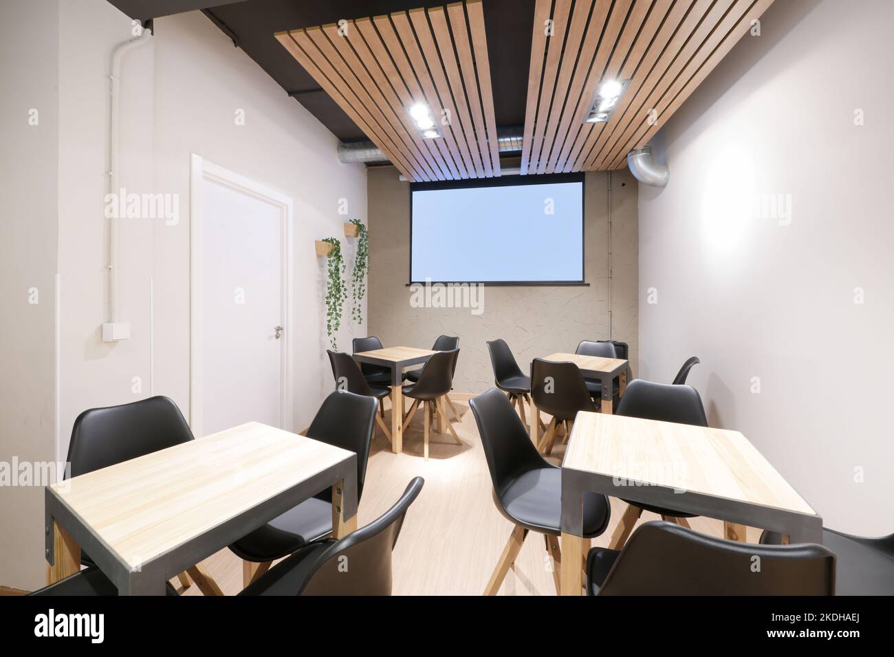 Salle à manger d'un restaurant avec des tables en bois de hêtre carré et en métal gris et un écran de projection sur un mur Banque D'Images