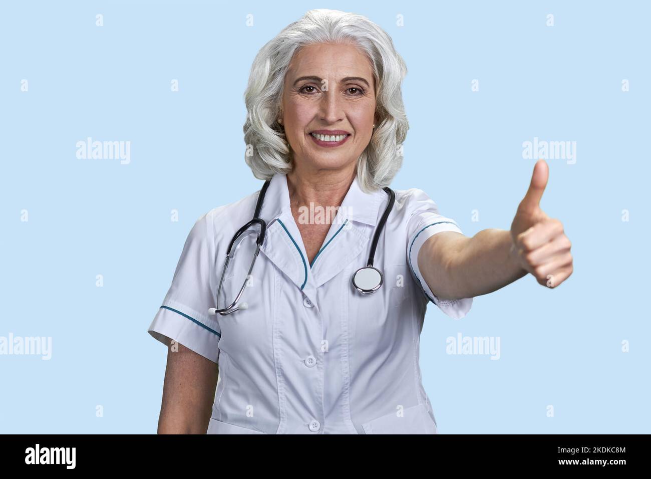 Portrait d'une femme âgée médecin montrant le pouce vers le haut. Femme âgée souriante avec un geste semblable. Fond bleu pâle. Banque D'Images