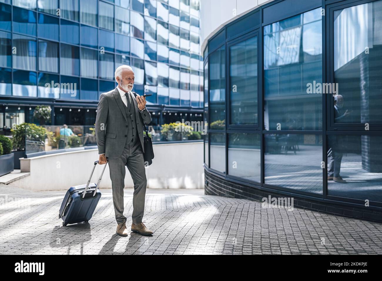 Homme âgé professionnel avec valise parlant à travers haut-parleur sur téléphone portable. Homme d'affaires senior vêtu d'un costume qui marche sur le sentier contre le bobu de bureau Banque D'Images