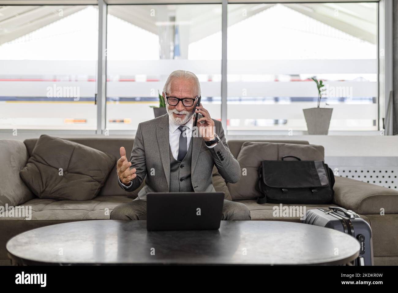 Un entrepreneur senior souriant parle sur un smartphone tout en étant assis sur un canapé. Un homme confiant regarde un ordinateur portable sur une table. Il porte pour Banque D'Images