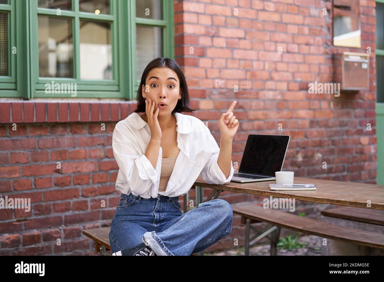 Élégante fille urbaine, femme asiatique avec ordinateur portable se trouve dans le café, points dans le coin supérieur droit bannière, montre l'espace de copie avec l'expression de visage surpris Banque D'Images