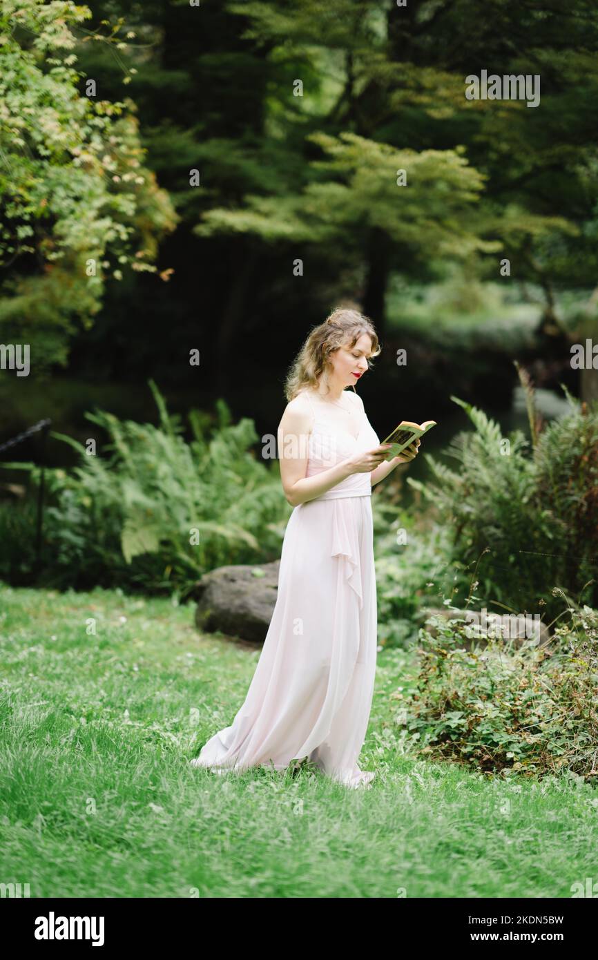 Femme portant un Gown rose lisant un livre dans un jardin idyllique Banque D'Images