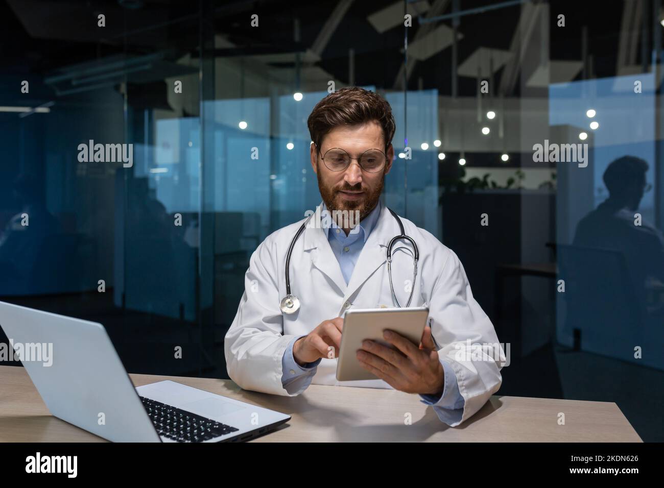 un médecin mature dans une robe médicale travaille à l'intérieur du bureau de la clinique, le médecin tient une tablette dans ses mains pour la consultation en ligne des patients, travaillant avec un ordinateur portable. Banque D'Images