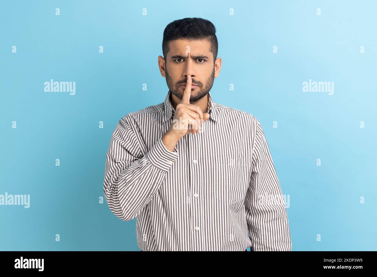 S'il vous plaît, Taisez-vous. Homme d'affaires tenant le doigt sur les lèvres faisant le geste de silence de hush, demandant de garder le secret, ne parlez pas, portant une chemise rayée. Studio d'intérieur isolé sur fond bleu. Banque D'Images