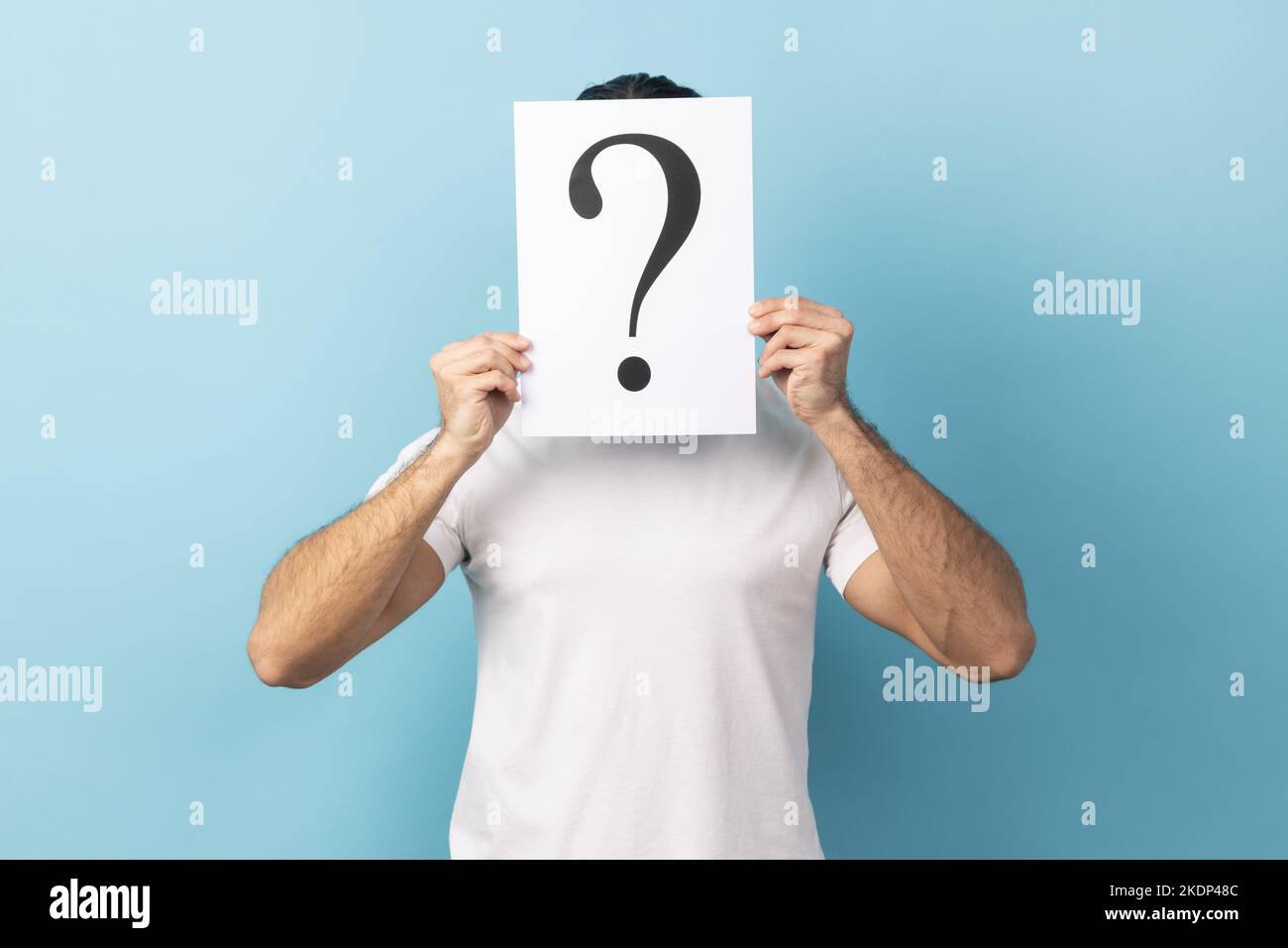 Portrait d'un homme portant un T-shirt blanc qui cache son visage derrière un livre blanc avec un point d'interrogation, qui recherche une solution intelligente et demande des conseils. Studio d'intérieur isolé sur fond bleu. Banque D'Images