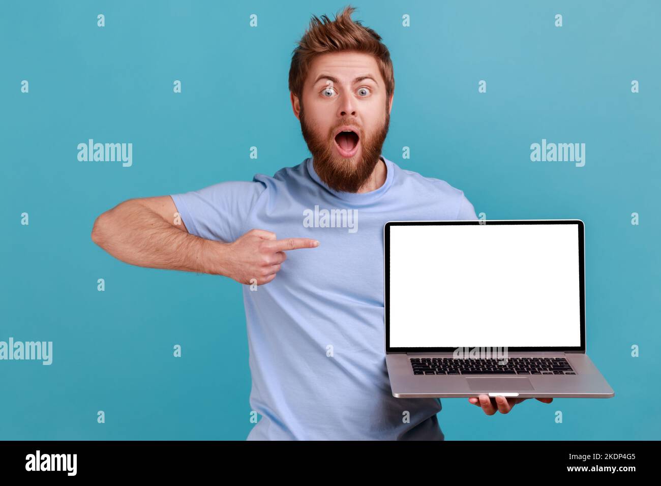 Portrait d'un homme barbu choqué tenant un ordinateur portable et pointant vers un écran vide, surpris par l'application informatique, regardant l'appareil photo. Studio d'intérieur isolé sur fond bleu. Banque D'Images